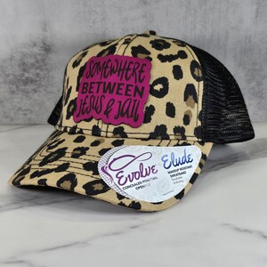 May include: A brown and black leopard print trucker hat with a pink patch that says "Somewhere Between Jesus & Jail". The hat has a black mesh back and a concealed ponytail opening.