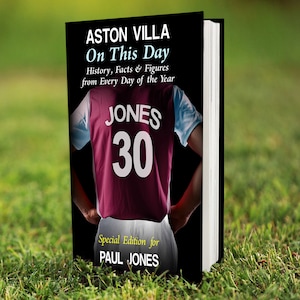 May include: A book titled "ASTON VILLA On This Day" with historical facts and figures. The cover shows a jersey with "JONES 30". The book is a special edition for "PAUL JONES". The book is set on a grassy background.
