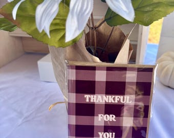 May include: A greeting card with a burgundy and cream plaid pattern. The card reads "THANKFUL FOR YOU" in white capital letters. The card is in front of a white flower arrangement with green leaves.