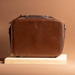 May include: A rectangular brown leather crossbody bag featuring a dark brown zippered top closure, visible stitching along the edges, and a small embossed fox head logo centered on the front panel. The bag includes metal hardware side rings.