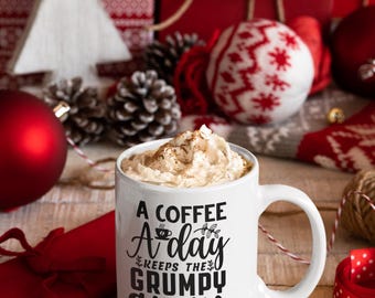 Op de afbeelding: Een witte keramische koffiemok met zwarte tekst die "A coffee a day keeps the grumpy away" luidt. De mok is gevuld met koffie en bedekt met slagroom. De mok is omringd door kerstversiering, waaronder een rood-witte gebreide sok, dennenappels en rood-wit lint.