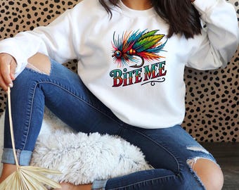May include: White sweatshirt featuring a colorful fishing lure graphic with the text "Bite Me." The graphic includes a detailed illustration of a fishing fly with vibrant feathers and a hook. The sweatshirt is a casual, long-sleeved garment.
