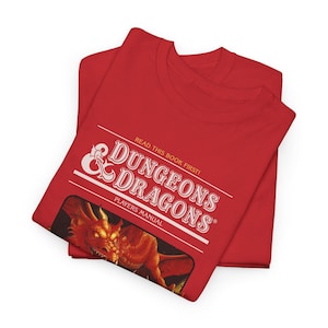 Op de afbeelding: Rood T-shirt met een wit Dungeons & Dragons logo en de tekst "Read this book first!" en "Players Manual". Het logo bevat een rode draak.