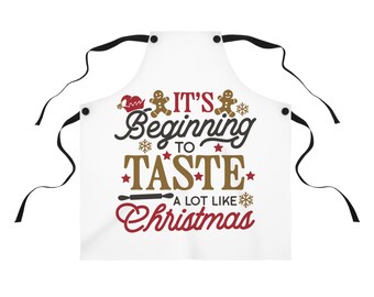 Puede incluir: Delantal blanco con lazos negros. El texto "It's Beginning to Taste a lot like Christmas" está impreso en marrón, rojo y negro. El diseño incluye hombres de jengibre, copos de nieve y un gorro de Papá Noel.