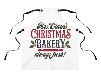 Könnte beinhalten: Weisse Schürze mit schwarzen Bindebändern. Auf der Schürze steht in roter und schwarzer Schrift "Mrs. Claus' CHRISTMAS BAKERY". Das Design umfasst Stechpalmenzweige und eine Nudelholz-Grafik mit dem Schriftzug "always fresh!" darunter.