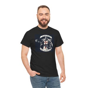 以下が含まれることがあります： 黒いTシャツ。胸に赤い十字架が付いた、メイスを持つ鎧姿の騎士のグラフィック。デザインには「BY LIGHT AND MIGHT」と「MORE THAN MERE HEALING」の文字が含まれています。