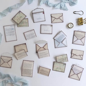 May include: A collection of vintage-style paper ephemera, including envelopes and handwritten letters. The items are in shades of beige and cream, with aged edges and a distressed appearance. A gold binder clip and a black paper clip are also visible.