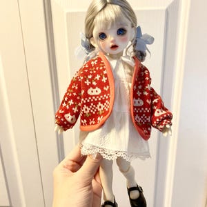 May include: A doll with blonde hair wearing a red and white patterned cardigan, a white dress with lace trim, and black shoes.