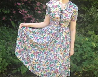 Könnte beinhalten: Ein Kleid mit Blumenmuster, kurzen Ärmeln und einem gerafften Bund. Das Kleid hat ein farbenfrohes Muster aus rosa, gelben und blauen Blumen auf weißem Grund. Ein schwarzes Band ist am Ausschnitt gebunden. Die Person steht auf grünem Gras.