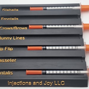 May include: A black organizer with gold lettering and several syringes with orange caps. The organizer has labeled slots for Glabella, Frontalis, Crows/Brows, Bunny Lines, Lip Flip, Masseter, and Mentalis. The text "Injections and Joy LLC" is at the bottom.