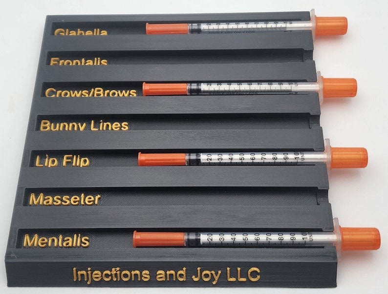 May include: A black organizer with gold lettering and several syringes with orange caps. The organizer has labeled slots for Glabella, Frontalis, Crows/Brows, Bunny Lines, Lip Flip, Masseter, and Mentalis. The text "Injections and Joy LLC" is at the bottom.