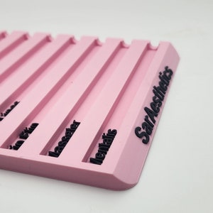 May include: A pink, rectangular product display with black text. The display has several parallel grooves and the words "SarAesthetics" are printed on the side. The display is on a white background.