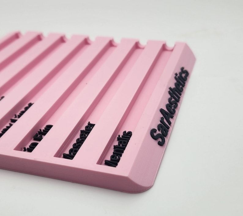 May include: A pink, rectangular product display with black text. The display has several parallel grooves and the words "SarAesthetics" are printed on the side. The display is on a white background.