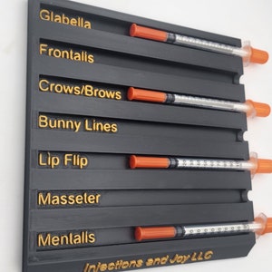 May include: A black organizer with gold lettering for cosmetic injections, labeled with terms like "Glabella" and "Bunny Lines." Each section holds a syringe with an orange cap. The text "Injections and Joy LLC" is at the bottom.