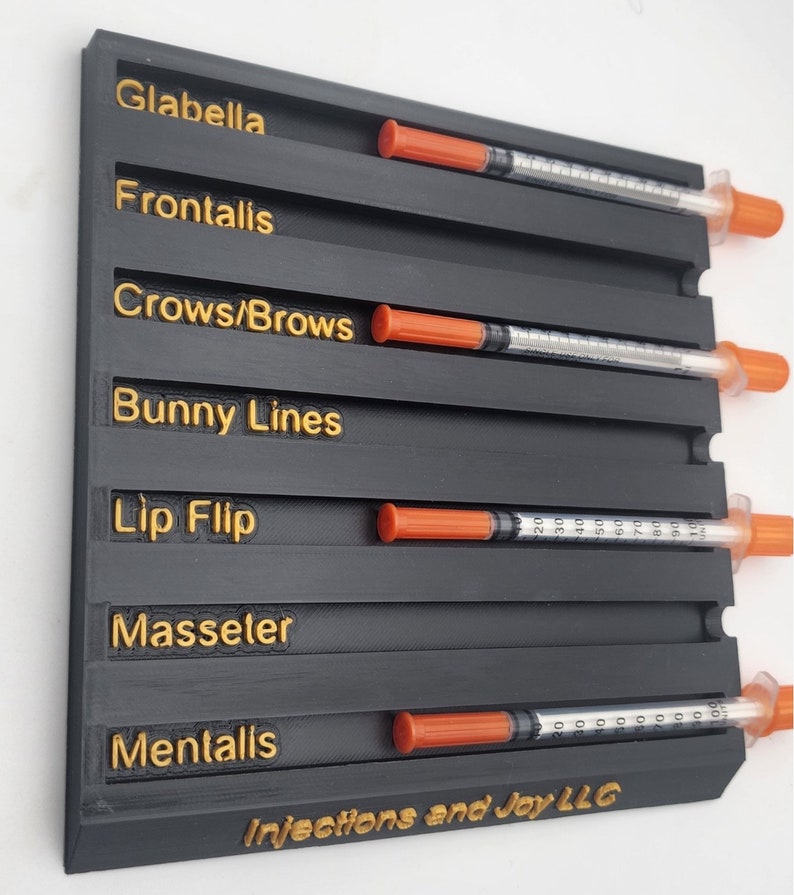 May include: A black organizer with gold lettering for cosmetic injections, labeled with terms like "Glabella" and "Bunny Lines." Each section holds a syringe with an orange cap. The text "Injections and Joy LLC" is at the bottom.