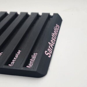 May include: A black, rectangular tray with multiple parallel grooves. The text "SarAesthetics" is printed in pink on the side. Additional pink text is visible, including the words "Masseter" and "Mentalis".