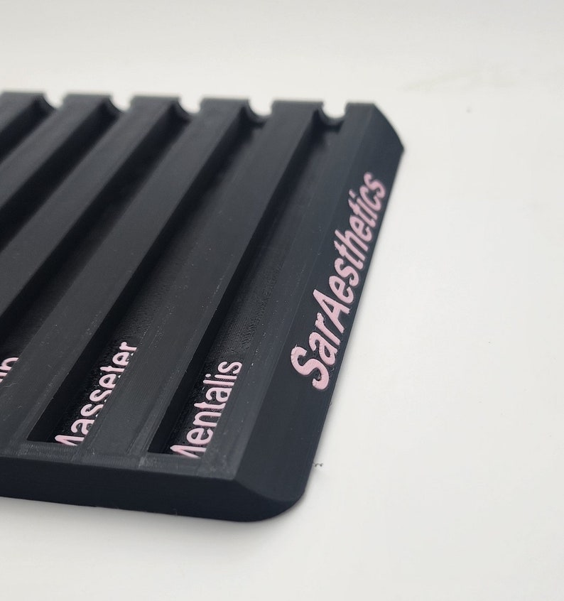 May include: A black, rectangular tray with multiple parallel grooves. The text "SarAesthetics" is printed in pink on the side. Additional pink text is visible, including the words "Masseter" and "Mentalis".