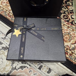 May include: A black gift box tied with a black ribbon printed with the words "SWEET" and "LOVE" in gold. A gold star charm is attached to the ribbon. The box rests on a patterned rug.