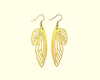 May include: A pair of gold-toned dangle earrings featuring a delicate, openwork insect wing design. Each earring is suspended from a thin gold-toned hook with a small clear plastic backing, showcasing intricate vein-like cutouts in the metal.