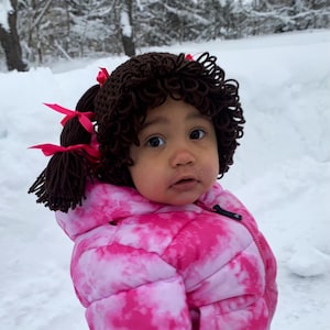 May include: A dark brown crocheted yarn wig featuring tight curls and two side ponytails, each accented with a bright pink ribbon bow. The wig is worn over a head, paired with a vibrant pink and white tie-dye patterned puffer jacket.