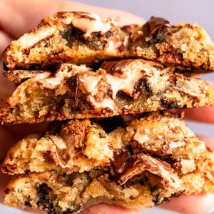 May include: A stack of four cookies, each cut in half to reveal the interior. The cookies are golden brown with visible chocolate chips and chunks, and a creamy, light-colored filling. The texture appears soft and chewy.