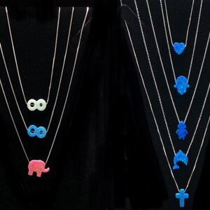 May include: Silver necklaces with opal pendants. One set features infinity symbols and an elephant, while the other showcases a heart, Hamsa hand, a figure, a dolphin, and a cross. The pendants are in shades of pink, blue, and white.