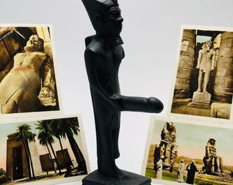 Peut inclure: Statue noire d'un pharaon égyptien avec un phallus proéminent, sur une base rectangulaire. Des cartes postales vintage de monuments égyptiens entourent la statue. La statue est d'un noir mat, évoquant l'art égyptien ancien.