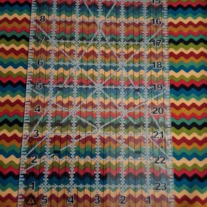 May include: A fabric featuring a repeating horizontal chevron pattern in shades of red, yellow, green, blue, and black. A clear plastic quilting ruler is placed vertically on top, showing measurements up to 15 inches. Eight small circular buttons are aligned at the bottom.