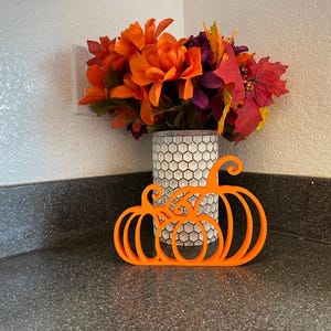 May include: A bright orange, flat, 3D-printed pumpkin silhouette decoration featuring three connected pumpkin shapes with curved stems. Behind the pumpkin cutout sits a cylindrical vase with a white and grey honeycomb pattern filled with artificial flowers.