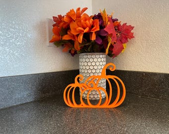 May include: A bright orange, flat, 3D-printed pumpkin silhouette decoration featuring three connected pumpkin shapes with curved stems. Behind the pumpkin cutout sits a cylindrical vase with a white and grey honeycomb pattern filled with artificial flowers.