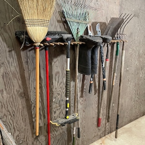 May include: A wooden wall rack with a variety of gardening tools hanging on it, including a broom, rake, shovel, and a squeegee. The tools are all in various shades of brown, green, and red.