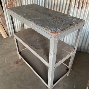 May include: A gray metal rolling cart with three shelves. The shelves are made of wood and have a worn, distressed look. The cart has four wheels and is perfect for storage or organization.