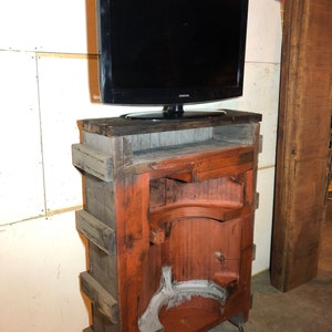May include: A rustic wooden TV stand with a distressed finish. The stand has a shelf for a TV and a lower shelf for storage. The stand is painted in shades of brown and red, with a gray interior. The stand has wheels for easy mobility.