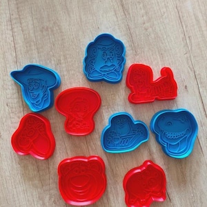 Peut inclure: Un ensemble de neuf emporte-pièces en plastique rouge et bleu, présentant des motifs de personnages en relief, notamment un cow-boy, un ranger de l'espace, un dinosaure, un cochon, un ours, un chien, une pomme de terre et d'autres visages.