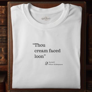 Puede incluir: Camiseta blanca con la cita "Thou cream faced loon" de Macbeth de William Shakespeare. La camiseta tiene cuello redondo y la marca "Quill & Thread Co" está impresa en la parte superior.