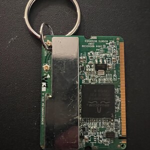 May include: A green circuit board with a silver metal cover and a silver key ring attached. The board has text that reads "2004 BROADCOM CORP ALL RIGHTS RESERVED 123" and "BCM94306MPLNA REV 4".