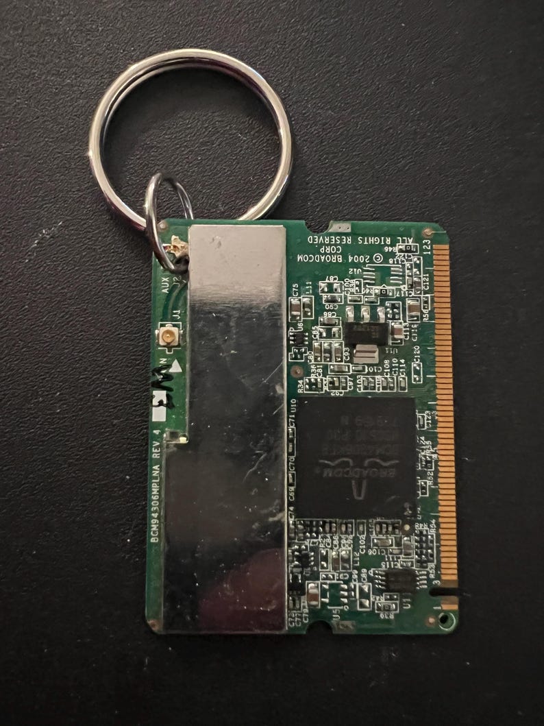 May include: A green circuit board with a silver metal cover and a silver key ring attached. The board has text that reads "2004 BROADCOM CORP ALL RIGHTS RESERVED 123" and "BCM94306MPLNA REV 4".