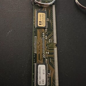 May include: A green circuit board keychain with a silver key ring. The circuit board has a black chip with the text "SEC KMM5324004ASW-6 KOREA 9729" and a smaller black chip with the text "hp 1818-6883 16MB EDO 60 (4Mx32) REORDER D3648-63001".