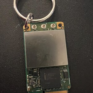 May include: A small green circuit board with a silver metal cover and three small buttons labeled 1, 2, and 3. The board has a gold connector on the bottom and is attached to a silver key ring.