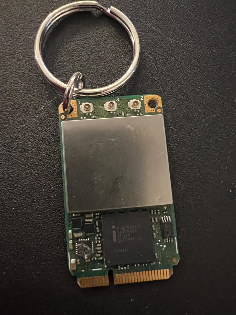May include: A small green circuit board with a silver metal cover and three small buttons labeled 1, 2, and 3. The board has a gold connector on the bottom and is attached to a silver key ring.