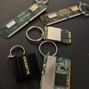 May include: Five keychains featuring different computer components. The keychains are made from old computer parts, including a black heatsink with the word "GIGABYTE" in gold lettering, a green circuit board with a silver metal cover, and a green circuit board with a silver metal cover.