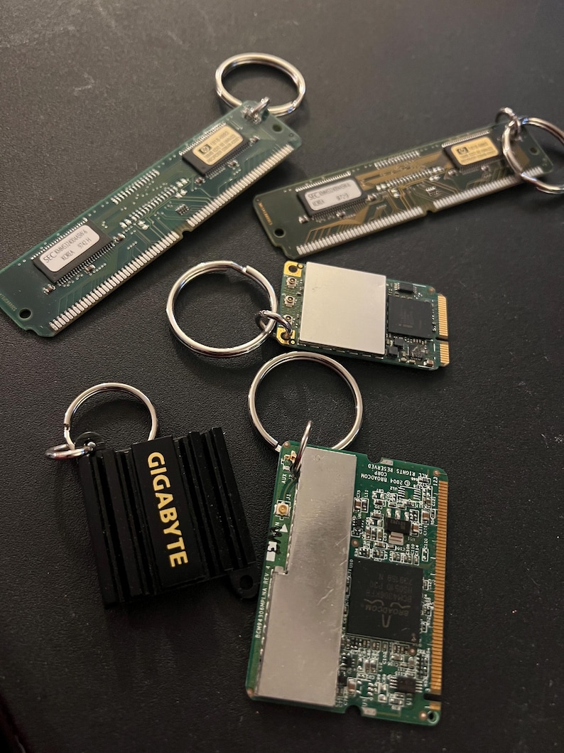 May include: Five keychains featuring different computer components. The keychains are made from old computer parts, including a black heatsink with the word "GIGABYTE" in gold lettering, a green circuit board with a silver metal cover, and a green circuit board with a silver metal cover.