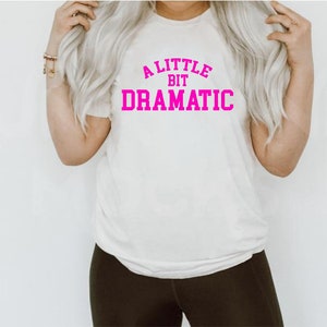 May include: White t-shirt with pink text that reads "A little bit dramatic".