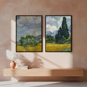 May include: Two framed landscape art prints. The artwork features a yellow field, green trees, and a cloudy sky. The frames are black. The artwork is displayed above a wooden shelf with a vase and decorative plant.