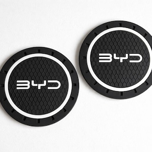 May include: Two black round car coasters with a white border and the letters "BYD" in white. The coasters have a textured surface and are set against a white background. Automotive accessory.
