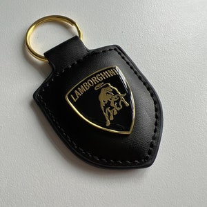 May include: Black leather keychain with a gold-colored Lamborghini emblem. The emblem features a gold bull and the word "Lamborghini". The keychain has a gold-colored ring.