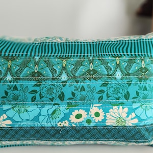 May include: A rectangular decorative pillow with a patchwork design. The pillow features horizontal bands of teal fabric with floral, butterfly, and striped patterns. The pillow is on a white surface.