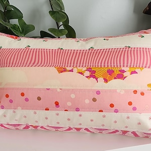 Op de afbeelding: Een rechthoekig decoratief kussen met een patchwork-ontwerp. Het kussen heeft horizontale strepen van verschillende roze en witte stoffen met patronen, waaronder strepen, stippen en bloemenprints. Het kussen ligt op een wit oppervlak.