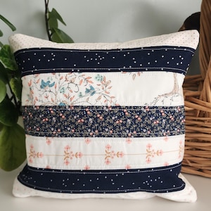 Peut inclure: Un coussin décoratif avec un motif patchwork. Le coussin présente des rayures alternées de tissus floraux et à motifs dans des tons marine, blanc et rose. Le coussin mesure environ 36 cm de large et 25 cm de haut.