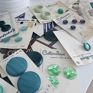 May include: A collection of vintage buttons in various shades of blue and green, displayed on their original cards. The buttons vary in size and design, with some featuring a marbled effect. The cards have text such as "Costumakers" and "Exquisit".